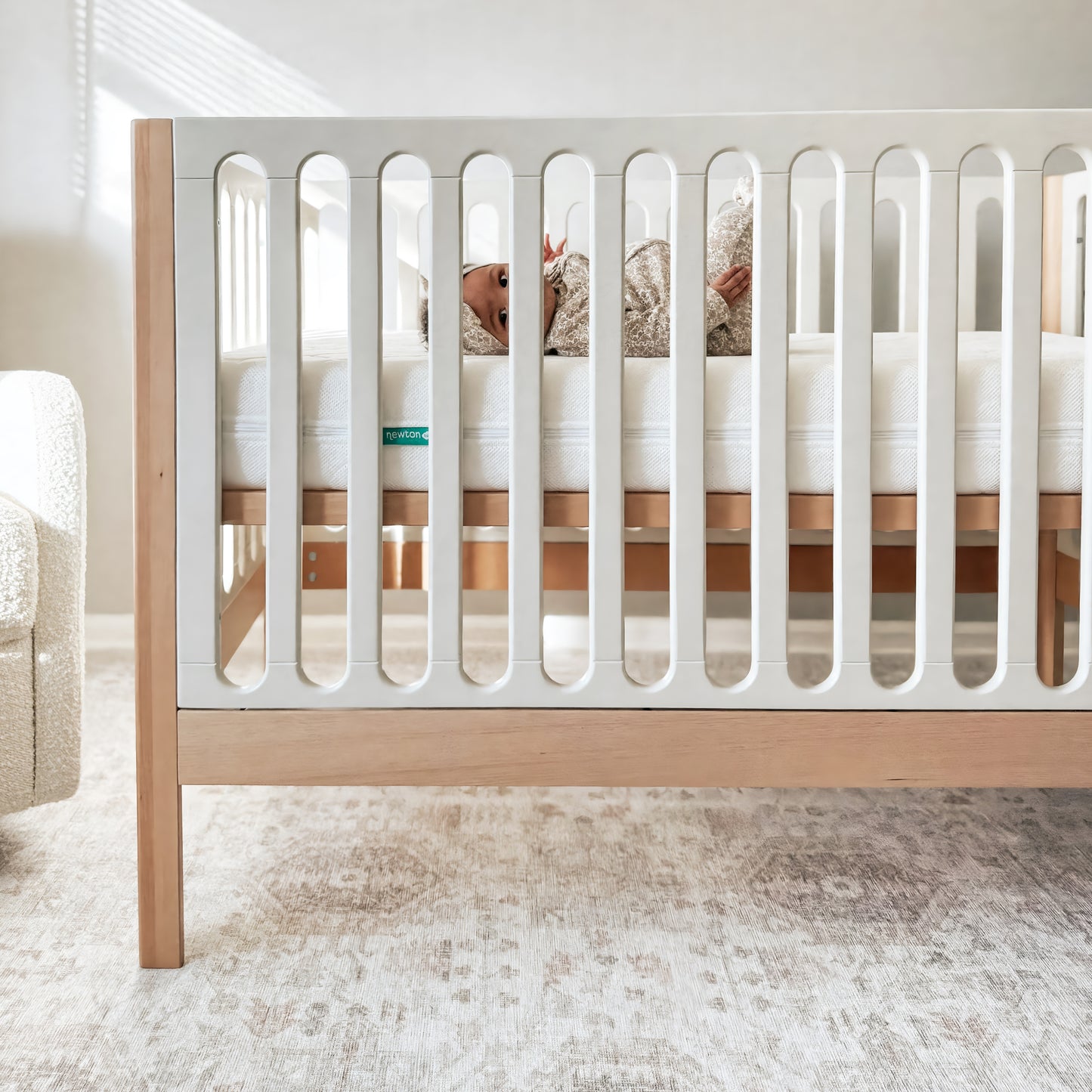 A baby rests in the Newton Galileo Convertible Crib by Newton Baby, crafted from white and beech wood. The softly lit room has neutral walls and carpet, with a white chair partially visible beside the crib.