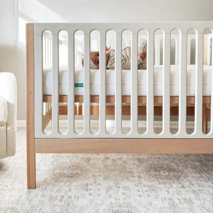 A baby rests in the Newton Galileo Convertible Crib by Newton Baby, crafted from white and beech wood. The softly lit room has neutral walls and carpet, with a white chair partially visible beside the crib.