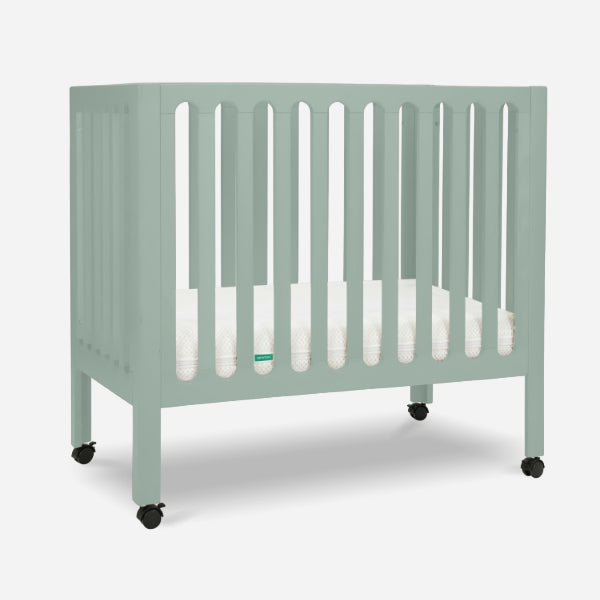 The Newton Baby Galileo Mini Crib in mint green features vertical slats, a white Newton mattress, and four black caster wheels, all set against a plain white background.