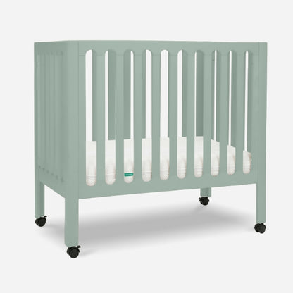 The Newton Baby Galileo Mini Crib in mint green features vertical slats, a white Newton mattress, and four black caster wheels, all set against a plain white background.