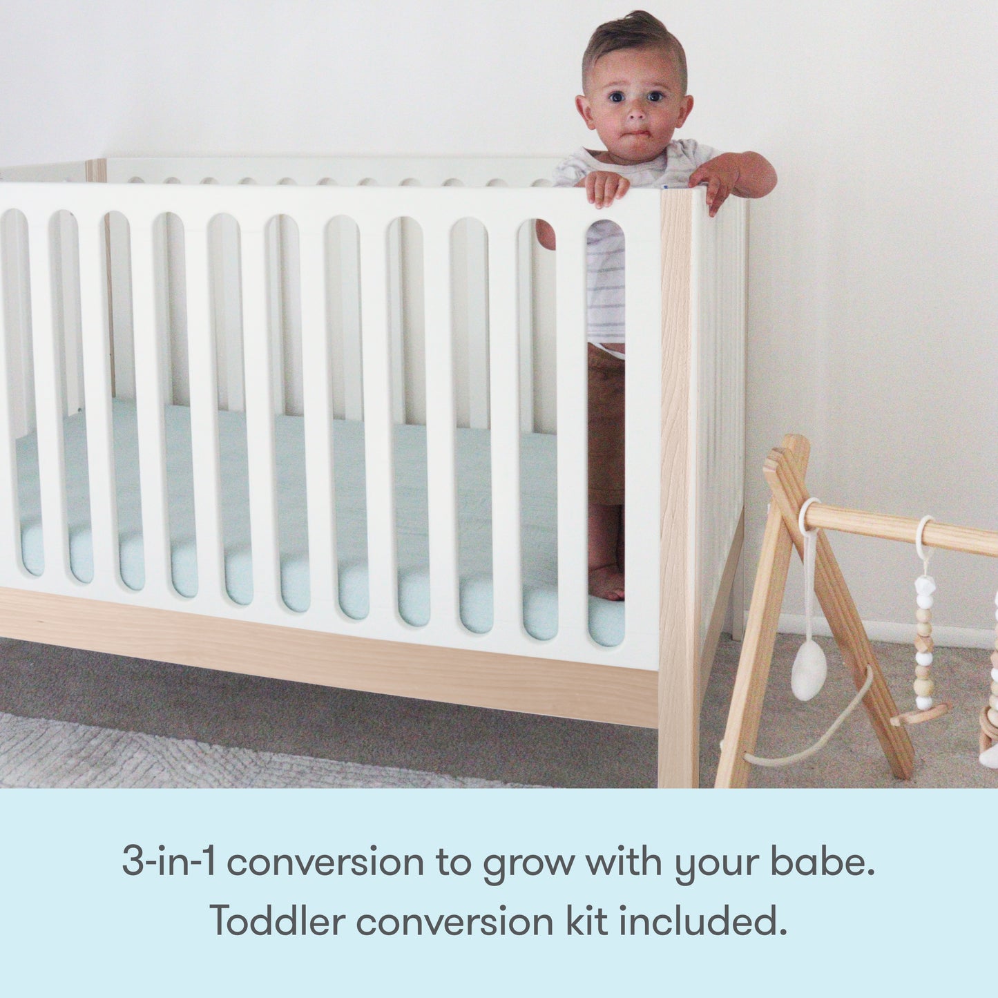A baby stands in a Newton Galileo Convertible Crib by Newton Baby, made of modern beech wood with vertical slats. A wooden baby gym with hanging toys sits nearby. Text: 3-in-1 crib grows with your baby—toddler bed conversion kit included.