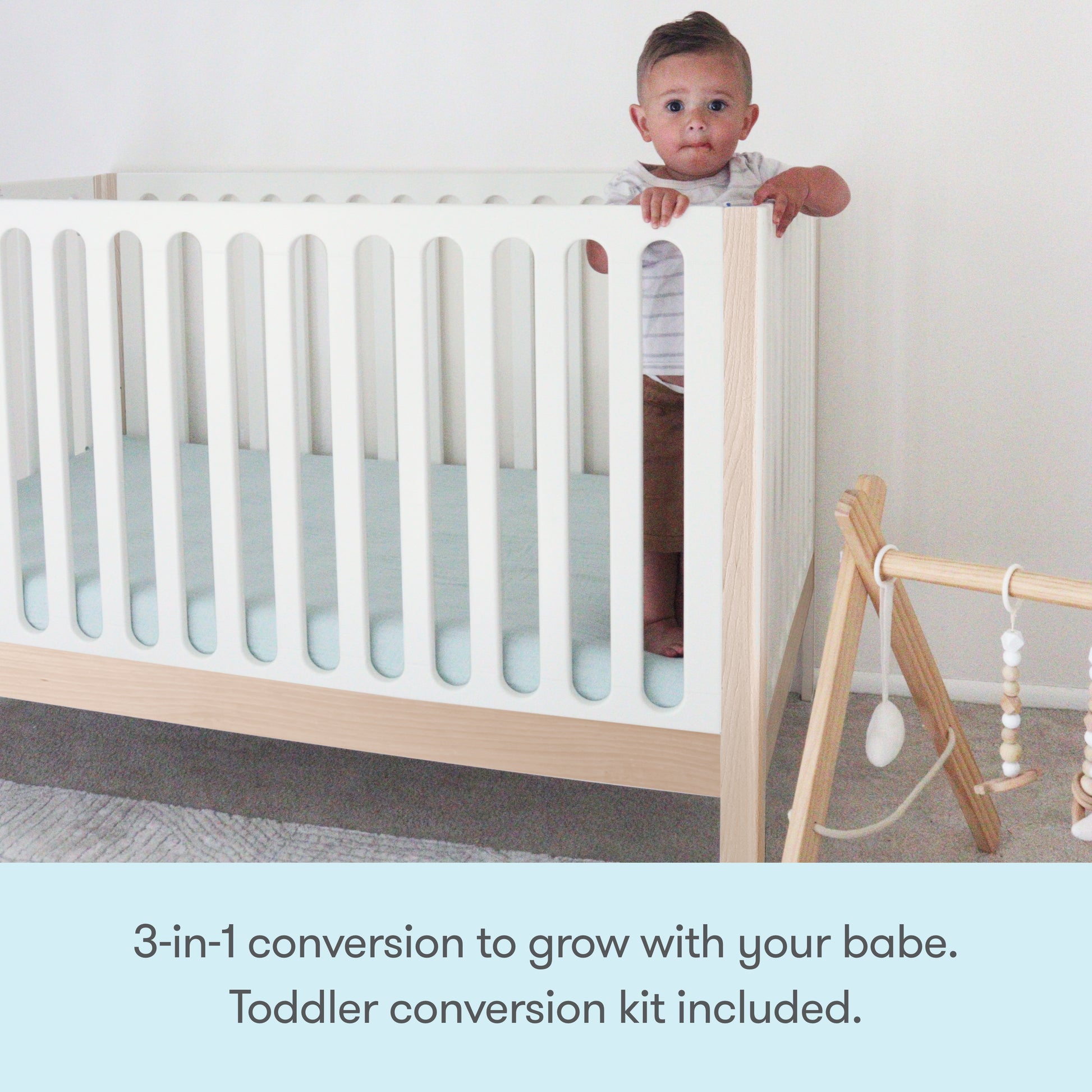 A baby stands in a Newton Galileo Convertible Crib by Newton Baby, made of modern beech wood with vertical slats. A wooden baby gym with hanging toys sits nearby. Text: 3-in-1 crib grows with your baby—toddler bed conversion kit included.