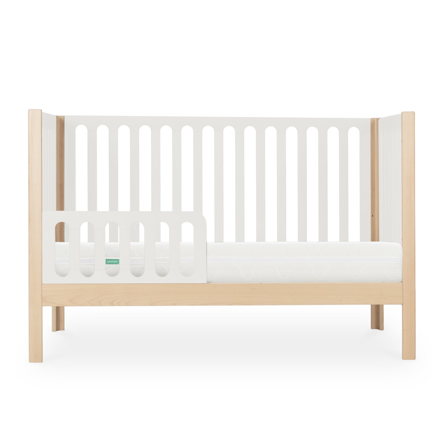 The Newton Galileo Convertible Crib by Newton Baby features natural beech wood legs, white slatted sides, and a low guard rail. Shown in toddler bed mode with a crib mattress, it’s set against a plain white background.