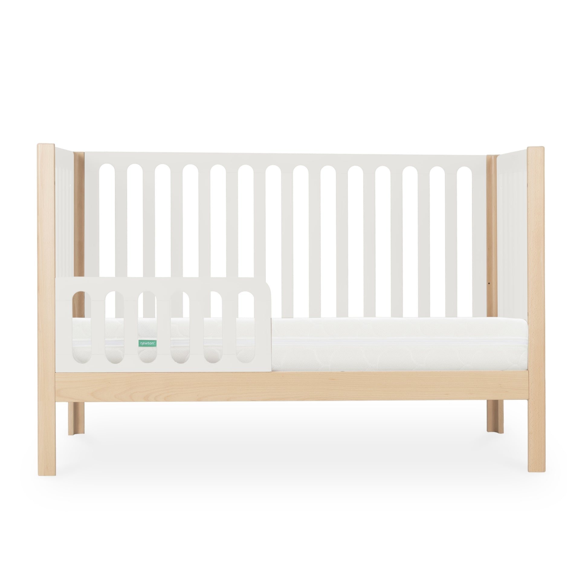 The Newton Galileo Convertible Crib by Newton Baby features natural beech wood legs, white slatted sides, and a low guard rail. Shown in toddler bed mode with a crib mattress, it’s set against a plain white background.