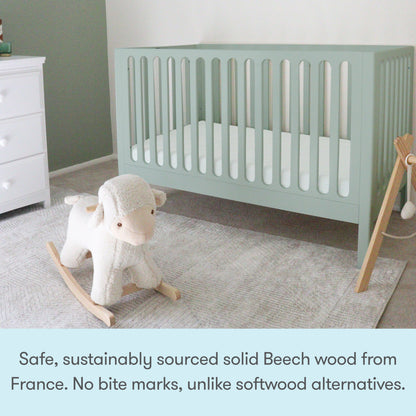 A light green Newton Galileo Convertible Crib by Newton Baby stands in a nursery with a plush rocking sheep on a soft grey rug. A white dresser is on the left. Text below highlights the crib’s safety features and sustainable materials.