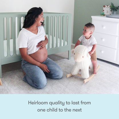 A pregnant woman kneels, smiling at a toddler on a rocking sheep beside the sleek Newton Galileo Convertible Crib by Newton Baby. Text below reads, Heirloom quality to last from one child to the next.