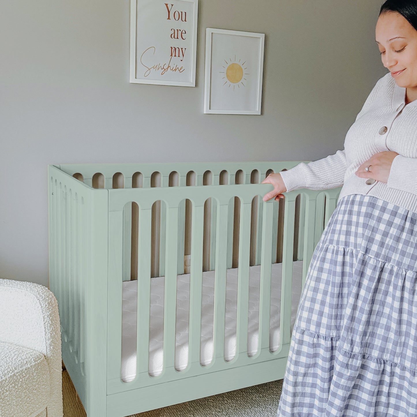 A pregnant woman stands beside the Newton Galileo Convertible Crib by Newton Baby in a softly lit nursery, gently touching her belly. Wall art features a sun and the phrase "You are my sunshine.