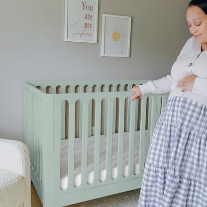A pregnant woman stands beside the Newton Galileo Convertible Crib by Newton Baby in a softly lit nursery, gently touching her belly. Wall art features a sun and the phrase "You are my sunshine.