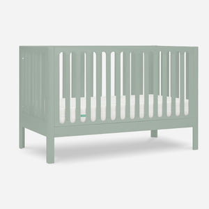The Newton Galileo Convertible Crib by Newton Baby features a light green finish, vertical slats on all sides, and includes a white mattress. Shown against a plain white background. Toddler bed conversion kit included for long-term use.