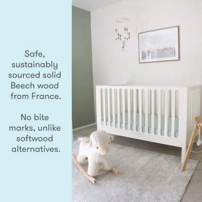 A baby's nursery featuring a white crib made from sustainably sourced solid Beech wood from France, accompanied by a text stating "Safe, sustainably sourced solid Beech wood from France. No bite marks, unlike softwood alternatives," with a toy sheep on a rocking base in the foreground. 