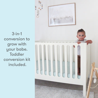 Toddler peeking over the side of a white 3-in-1 convertible crib, with text stating "3-in-1 conversion to grow with your babe. Toddler conversion kit included."