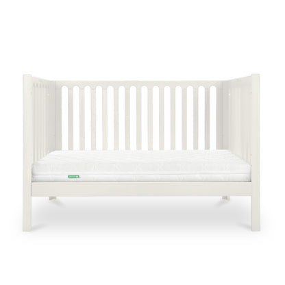 A simple white baby crib with a clean mattress displaying a small green 'Newton' logo on its corner.