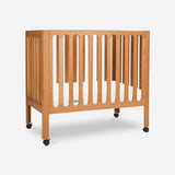 The Newton Baby Galileo Mini Crib with Newton Mattress is crafted from solid Beech wood, has vertical slats, a white mattress, and wheels on each leg for easy mobility, shown against a plain white background.