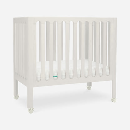 The Newton Baby Galileo Mini Crib with Newton Mattress, featuring vertical slats and four wheels, is crafted from solid Beech wood and shown on a plain white background.