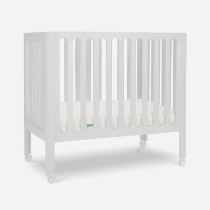 The Newton Galileo Mini Crib with Newton Mattress by Newton Baby features solid Beech wood, vertical slats on all sides, a thin mattress, and four wheels for easy mobility. The portable crib is shown against a plain white background.
