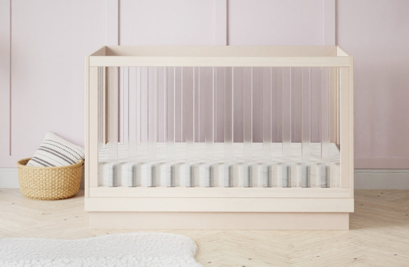 A modern wooden crib with a breathable mattress in a nursery room decorated with a pale pink wall and a basket beside it.