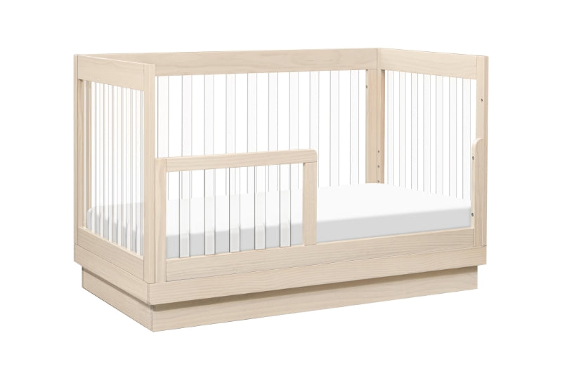 Modern wooden crib with a visible mattress, featuring vertical slats and a lower front gate for easy access.