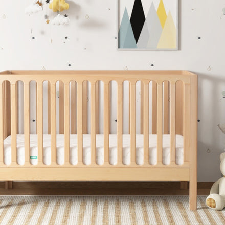 Light wood Newton crib centered in a nursery; teddy bear and basket fully visible on the right, with a partially visible white chair on the left.