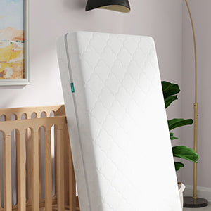 A white quilted mattress with delicate scalloped patterns leans against a light beige wooden crib. The crib's tall slats frame the background, while a teal label adds a subtle pop of color. Above, a soft watercolor landscape in warm tones hangs on a blush-gray wall, complemented by a sleek black pendant light. A lush green plant and gold floor lamp add organic warmth to the serene space.