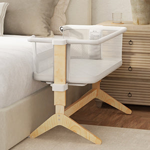 A light beige/cream-colored wooden crib, with a white mesh top, is positioned next to a bed. The crib has a simple, modern design with light beige/natural-colored wooden legs and a sturdy frame. The crib’s top is a white mesh, allowing for good air circulation, and the crib’s surface is also white. The crib appears to be attachable to a standard bed frame.  The room is light and neutral, with light beige/cream bedding and walls. A light colored wooden dresser or chest of drawers is visible in the background