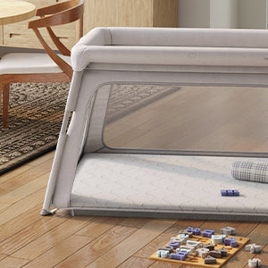 A light gray portable playpen sits on hardwood floors, featuring a padded top and breathable mesh sides. Inside, a cushioned gray-and-white patterned mattress provides a soft spot for rest. Nearby, wooden letters and numbers in muted purple and brown tones are playfully arranged on a small board. The scene is completed by a peek of a gray-cream rug and the legs of light wood dining chairs, adding warmth to the minimalist space.