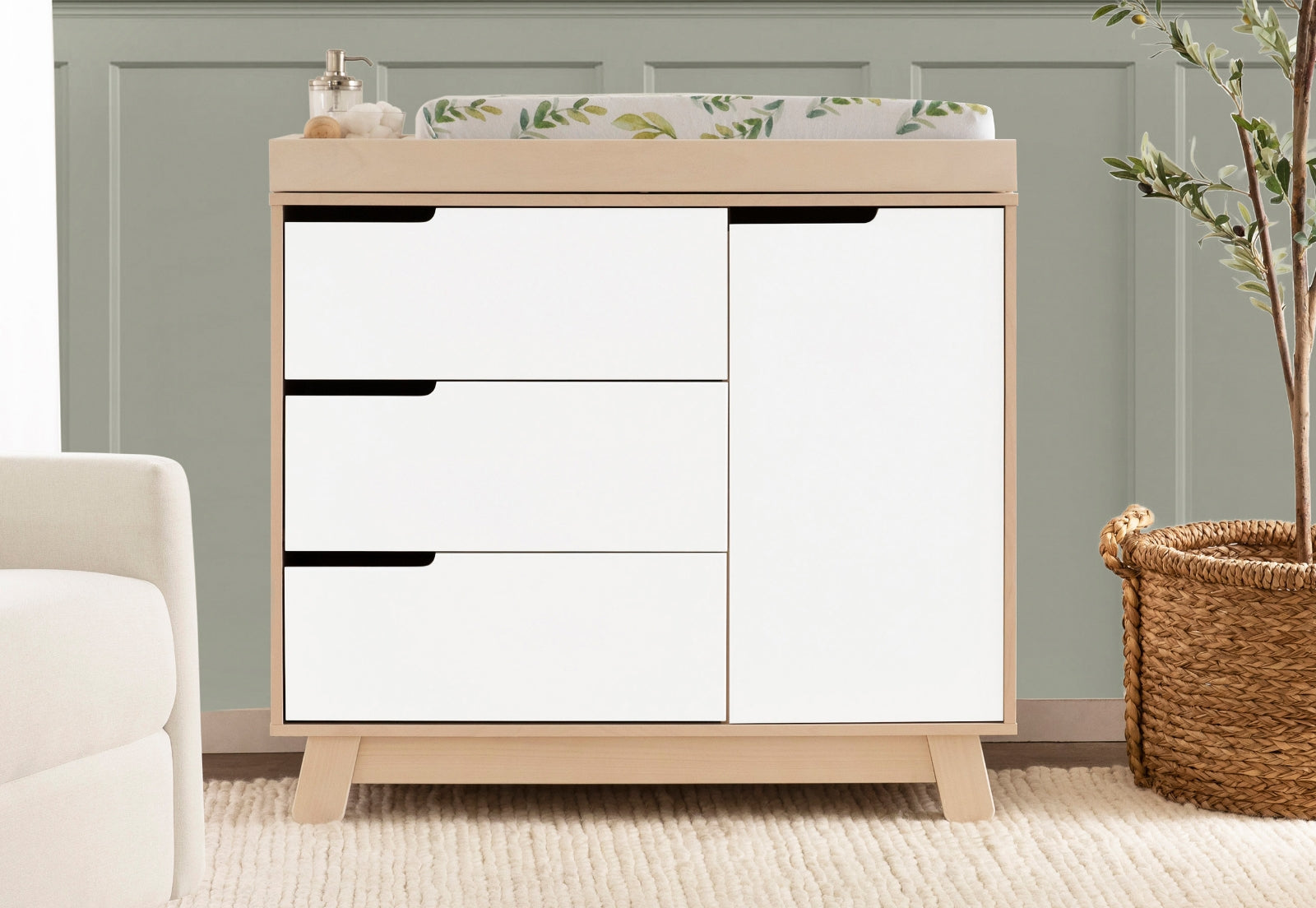 Modern nursery with a stylish white and wood changing table, featuring a plant and baby essentials on top, placed against a soft green wall. 999-8601-WWH