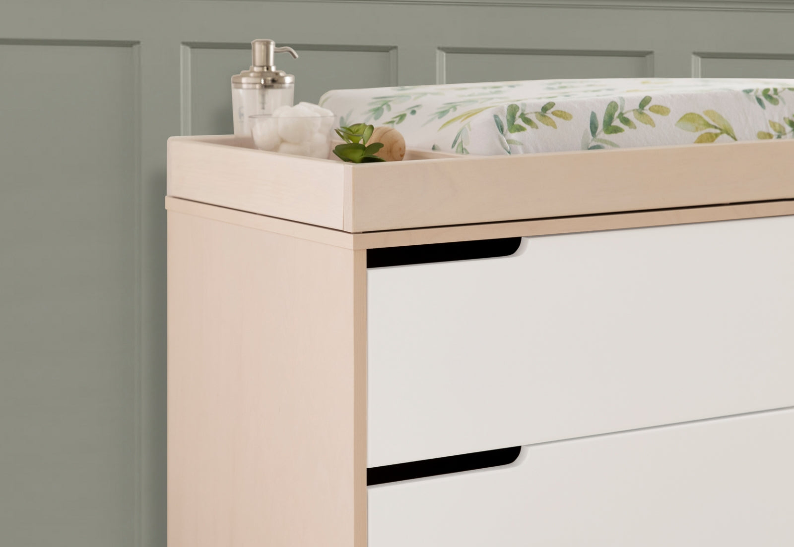 Modern nursery dresser in light wood finish featuring a changing pad with a leafy, green motif, accompanied by a dispenser and a small potted plant on top.