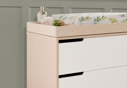 Modern nursery dresser in light wood finish featuring a changing pad with a leafy, green motif, accompanied by a dispenser and a small potted plant on top.