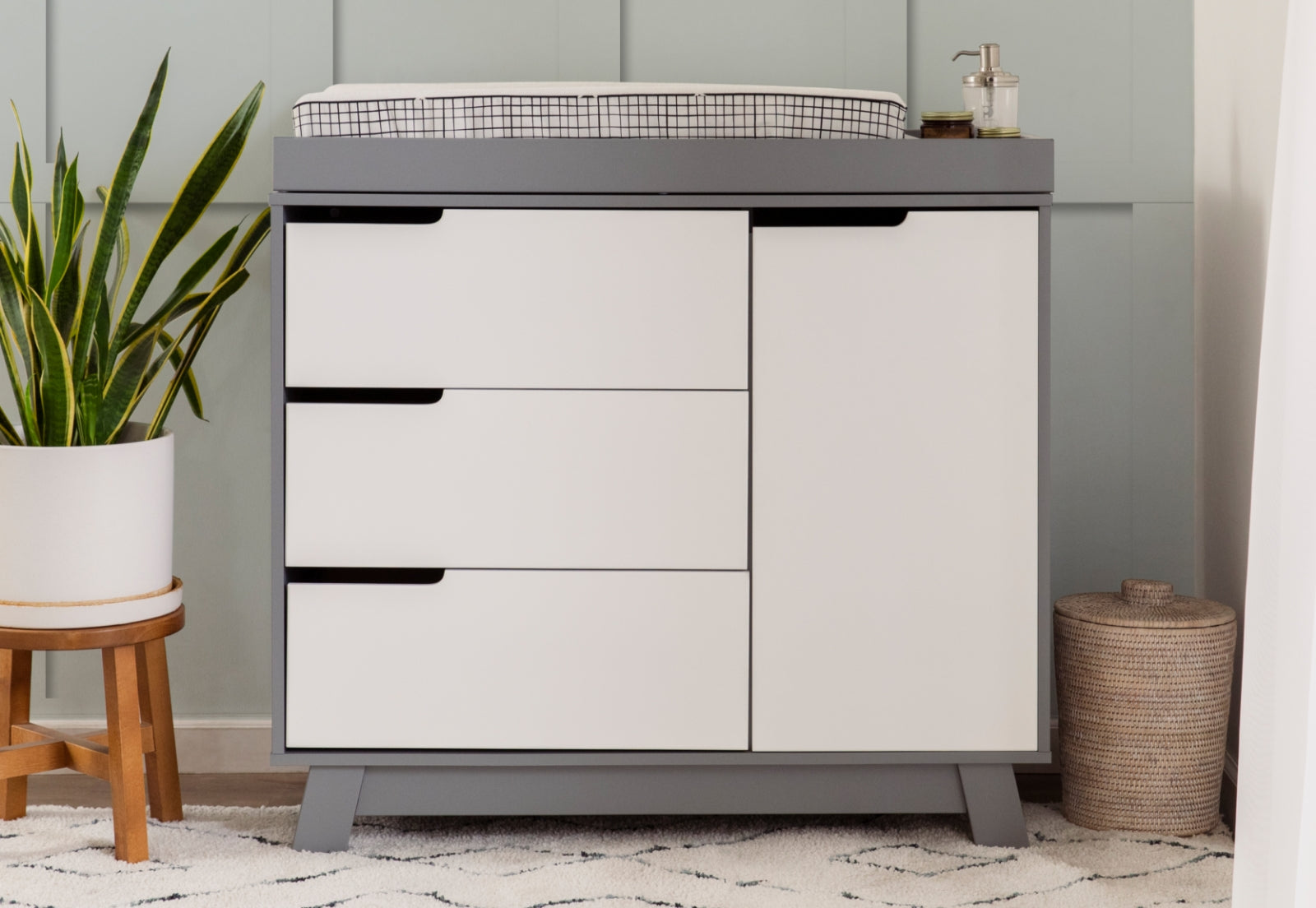 A modern changing table, set in a nursery featuring a plant and a woven basket, against a light-blue wall. 999-8601-GYW