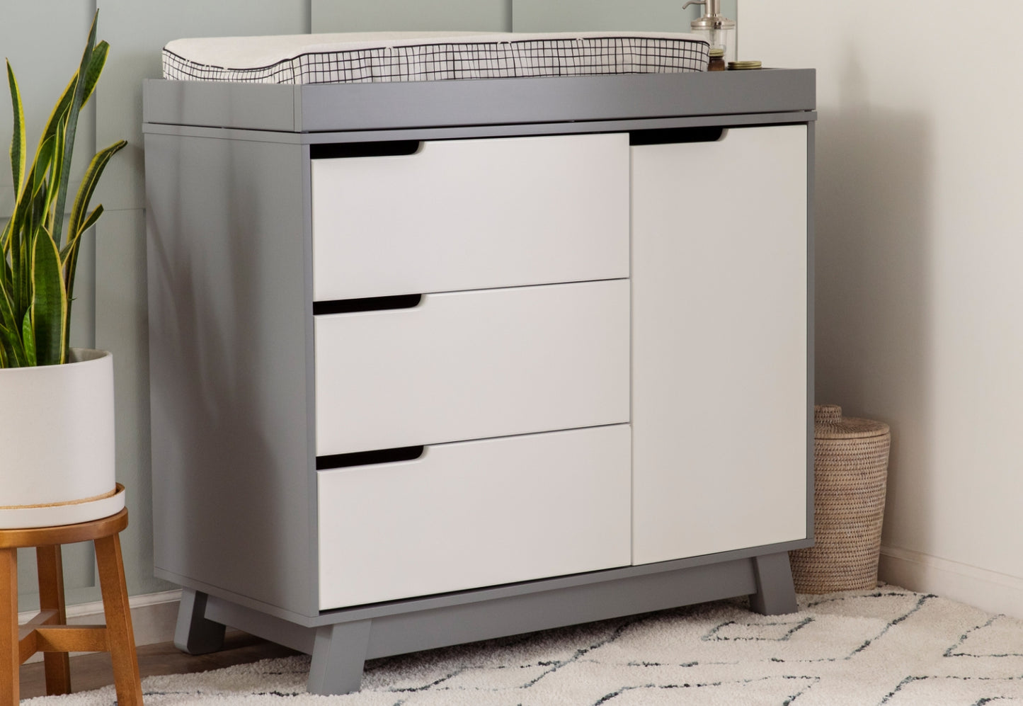 Modern gray and white nursery changing table with three drawers, topped with a changing pad, beside a plant and a basket.