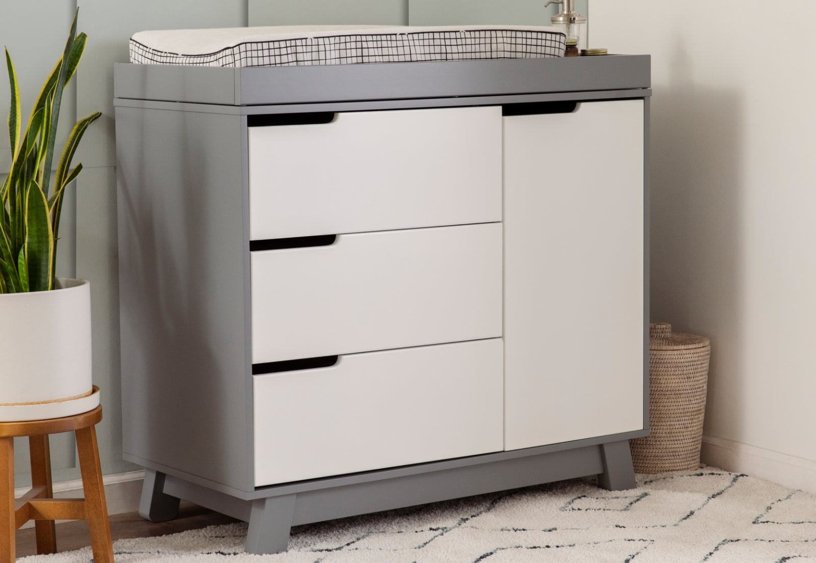 Modern gray and white nursery changing table with three drawers, topped with a changing pad, beside a plant and a basket.