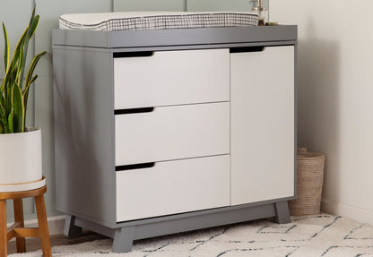 Modern gray and white nursery changing table with three drawers, topped with a changing pad, beside a plant and a basket.