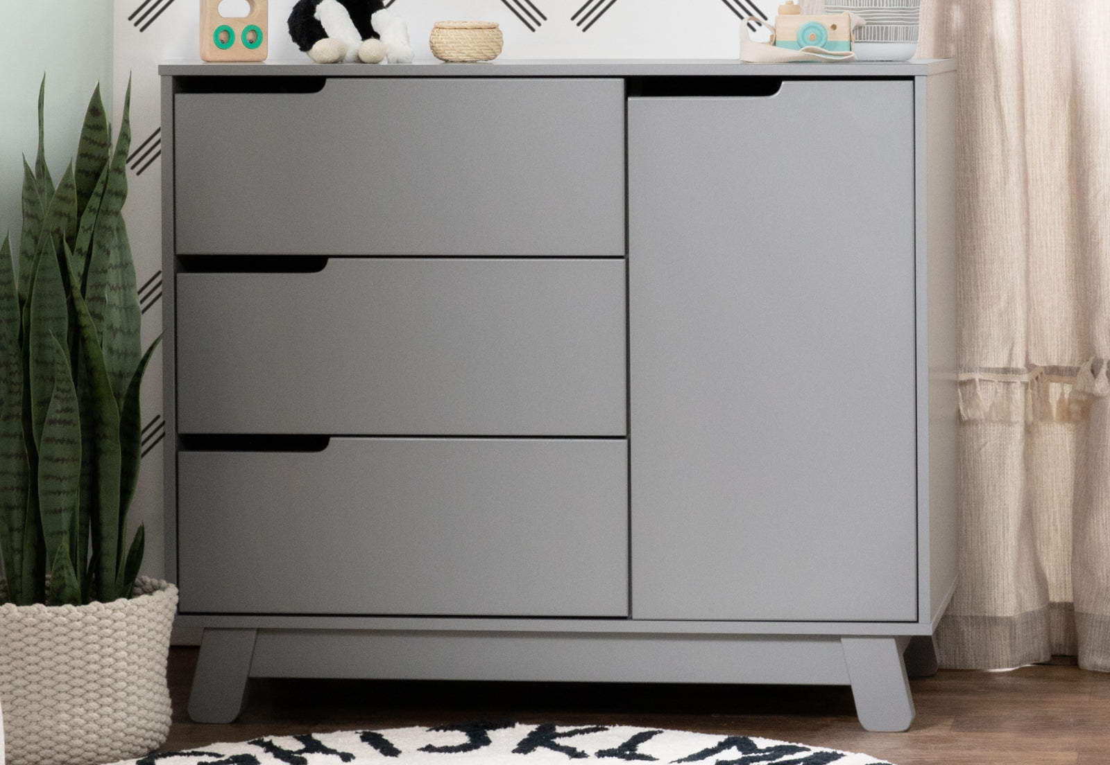 Modern gray nursery dresser with sleek lines and black handles, decorated with a small plant, toys, and a basket on top in a cozy room setting.