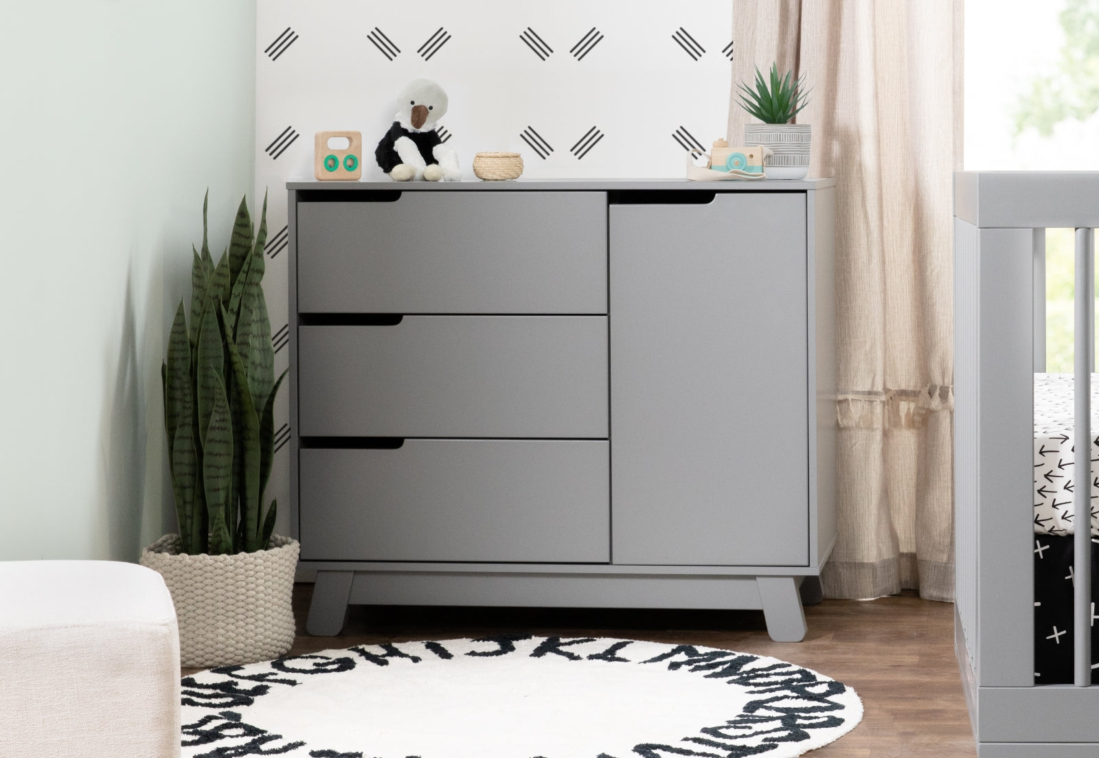 Modern gray nursery dresser with simple decorations, placed beside a crib, in a room with plant accents and a patterned rug.