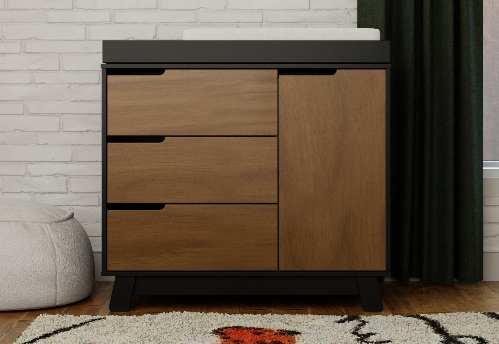 Modern baby changing table with a combination of walnut drawers and a solid door, featuring a black top and base, set against a white brick wall and a dark green curtain. 999-8601-NWB
