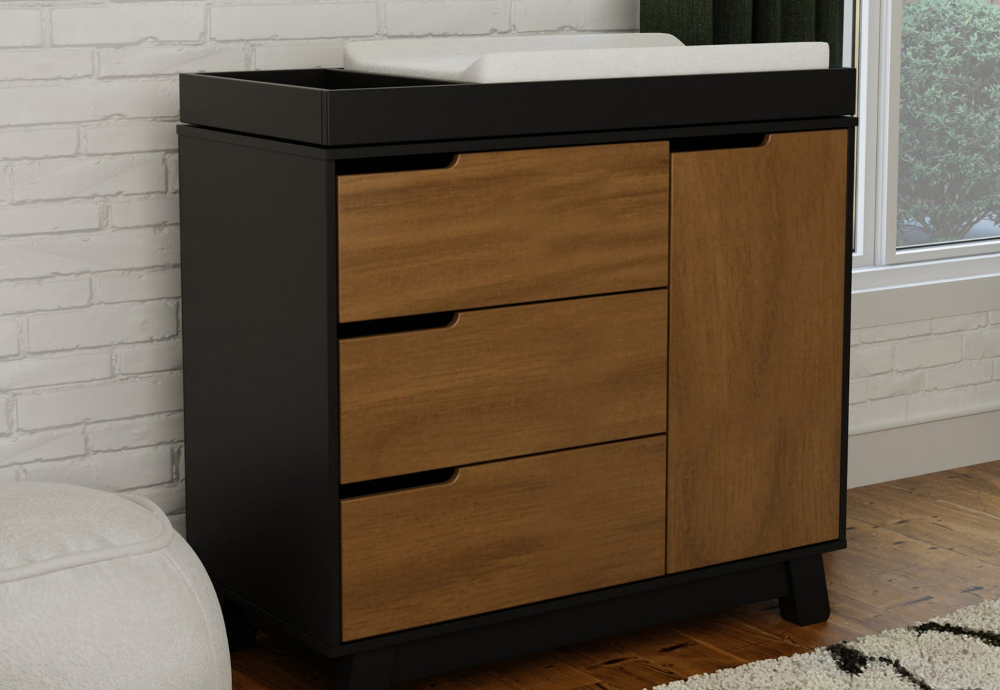 Stylish and functional nursery changing table with a dark base and natural wood drawers against a white brick wall.