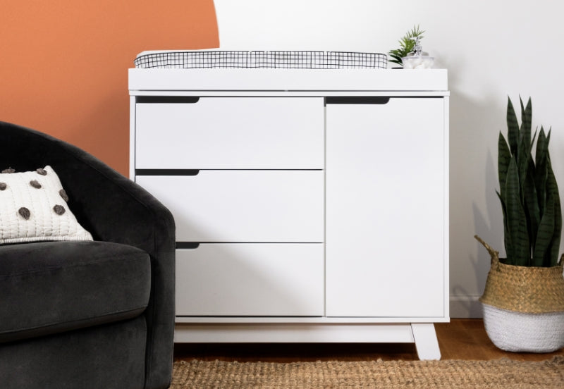 A modern white dresser with a black wire basket on top and a small plant, positioned beside a grey armchair and a potted snake plant in a contemporary styled room. 999-8601-WHT