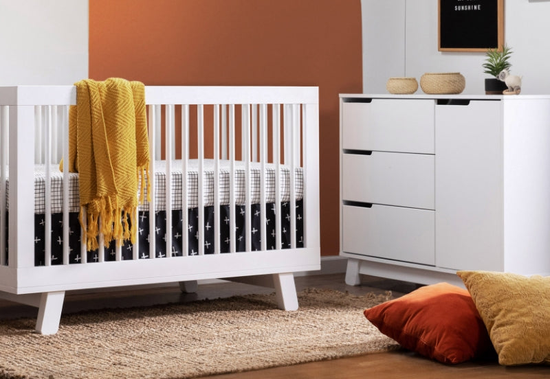 Modern nursery room featuring a white crib with a yellow blanket, adjacent to a white dresser, against a backdrop of orange and white walls, decorated with simple accessories.
