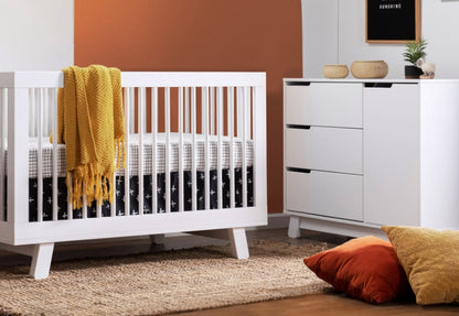 Modern nursery room featuring a white crib with a yellow blanket, adjacent to a white dresser, against a backdrop of orange and white walls, decorated with simple accessories.