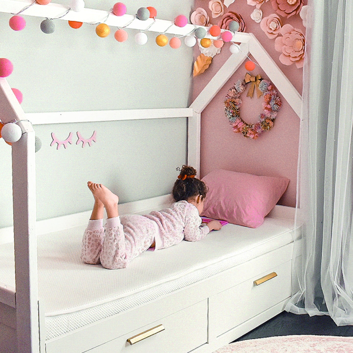 A young child in pajamas lies on a white house-shaped bed, watching a tablet. The pastel room features paper flowers, string lights, and a Newton Baby Kids' Twin Mattress that’s GREENGUARD Gold certified for safety.