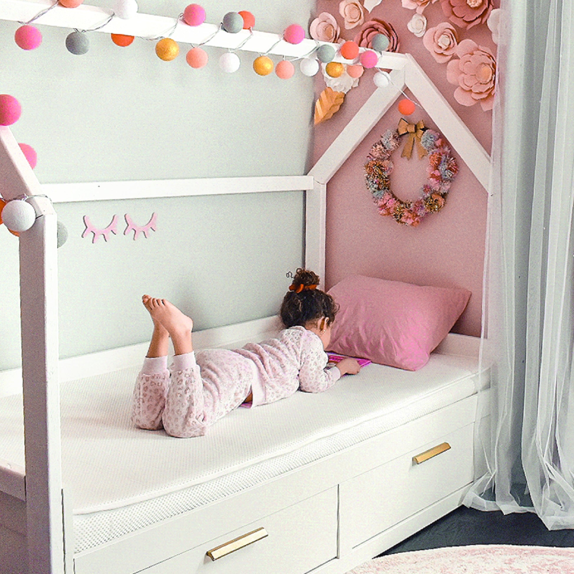 A young child in pajamas lies on a white house-shaped bed, watching a tablet. The pastel room features paper flowers, string lights, and a Newton Baby Kids' Twin Mattress that’s GREENGUARD Gold certified for safety.
