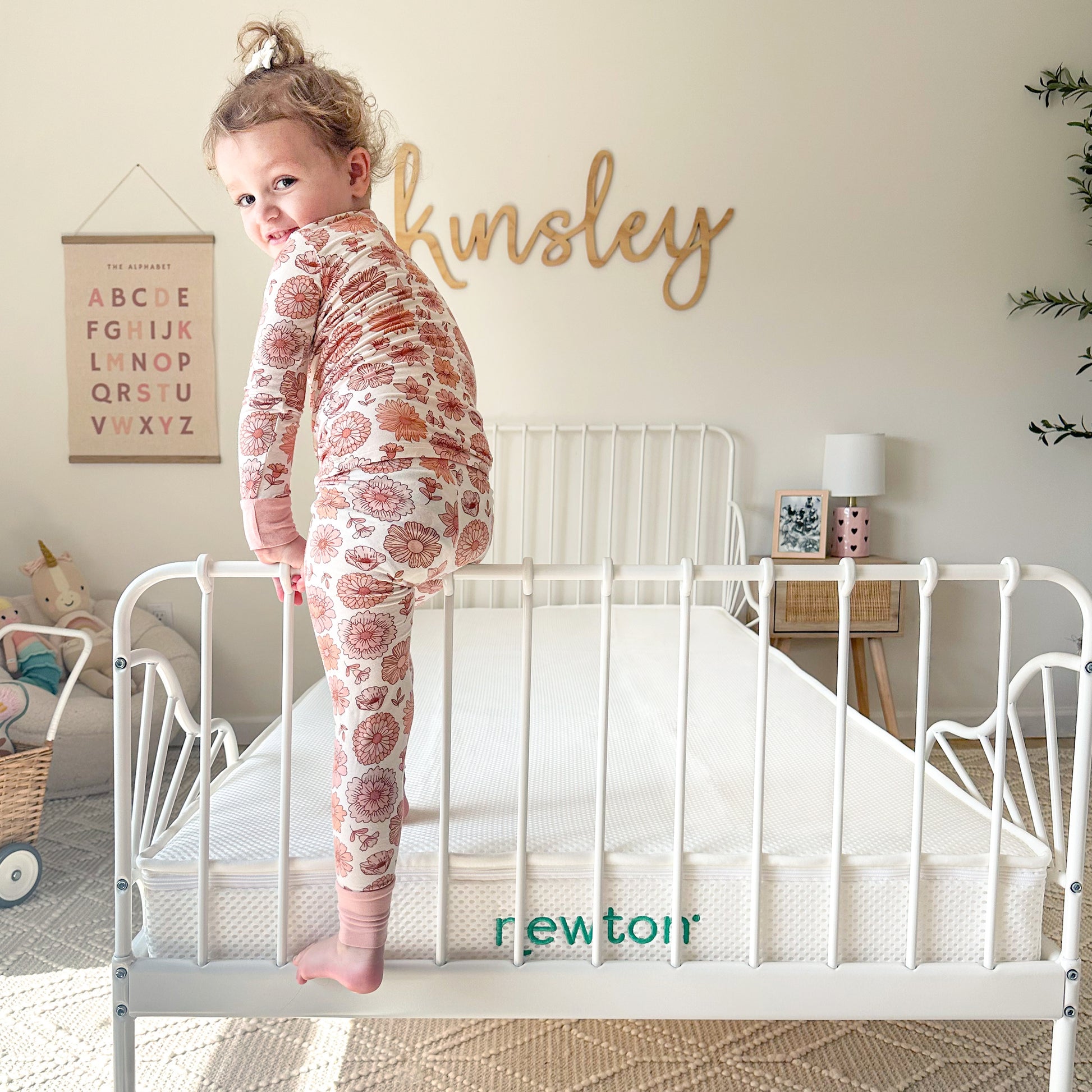A young child in patterned pajamas stands on a white metal bed frame with a breathable Kids' Twin Mattress by Newton Baby. The bright room features "Kinsley" on the wall and alphabet art above a toy basket.