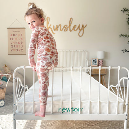 A young child in patterned pajamas stands on a white metal bed frame with a breathable Kids' Twin Mattress by Newton Baby. The bright room features "Kinsley" on the wall and alphabet art above a toy basket.