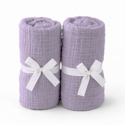 Two rolled, light purple towels with a textured pattern, each tied with a white ribbon bow and placed side by side on a plain white background—evoking the softness of Newton Baby’s Breathable, Organic Cotton Sheets (2-pack).