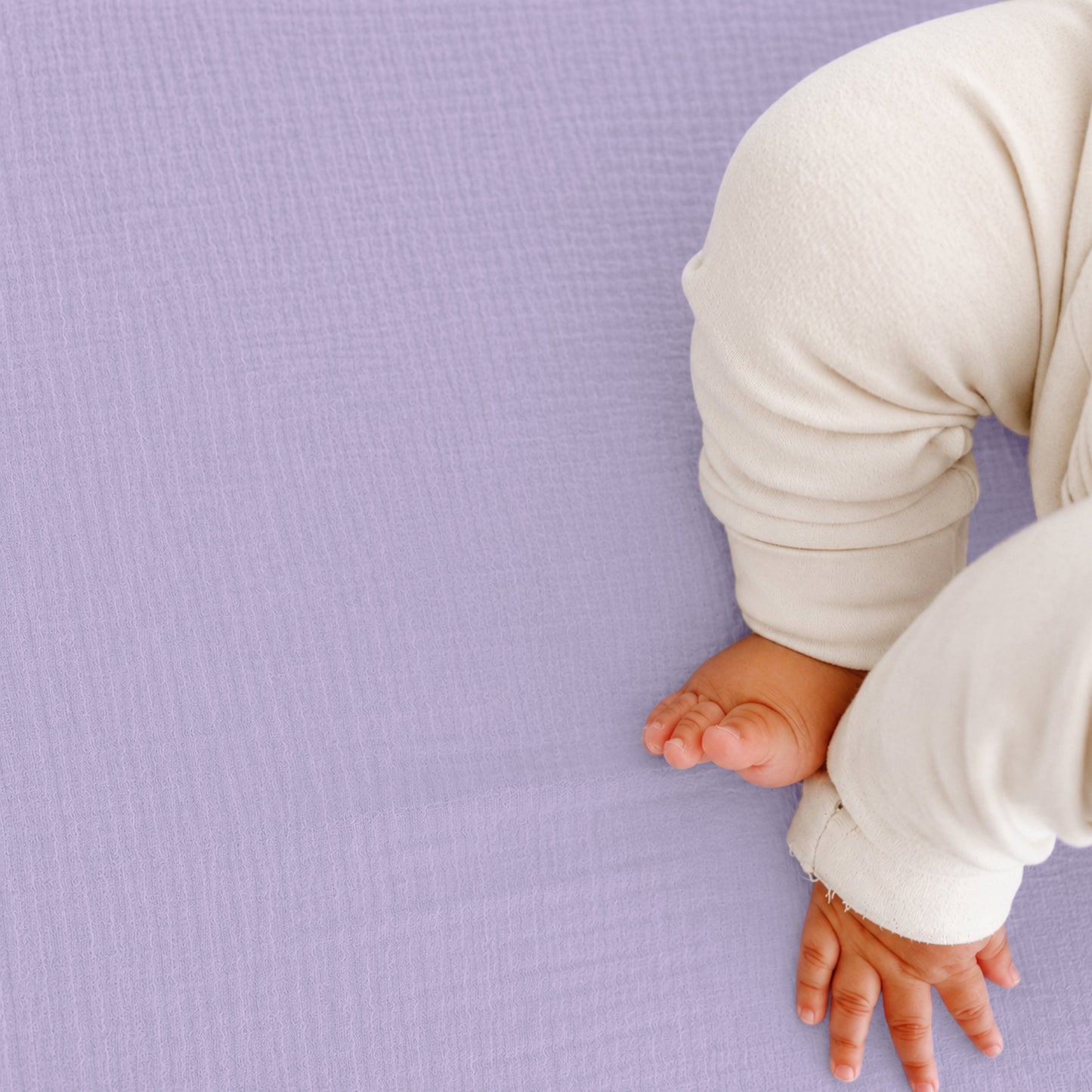 A baby, wearing a cream outfit, rests on a textured lavender surface—likely the Newton Baby Breathable, Organic Cotton Sheets (2-pack)—with only their arm, hand, and legs in view.