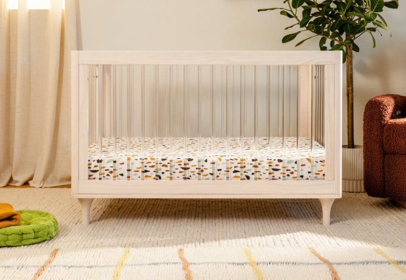 A modern white crib with a colorful mattress in a cozy room with a plush green pillow, patterned rug, and houseplants. 999-8502-WNA