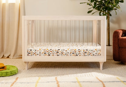 A modern white crib with a colorful mattress in a cozy room with a plush green pillow, patterned rug, and houseplants. 999-8502-WNA