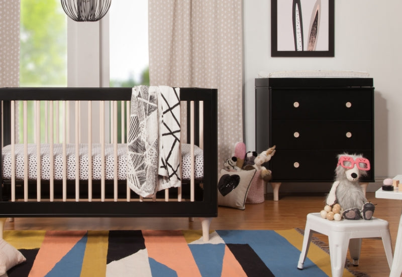 A whimsically styled nursery featuring a black crib with a patterned blanket, a dresser, a colorful rug, and a plush sloth wearing pink glasses sitting on a small white chair.