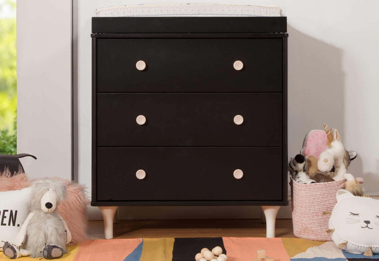 Modern nursery with a sleek black dresser flanked by playful toys and a colorful rug.