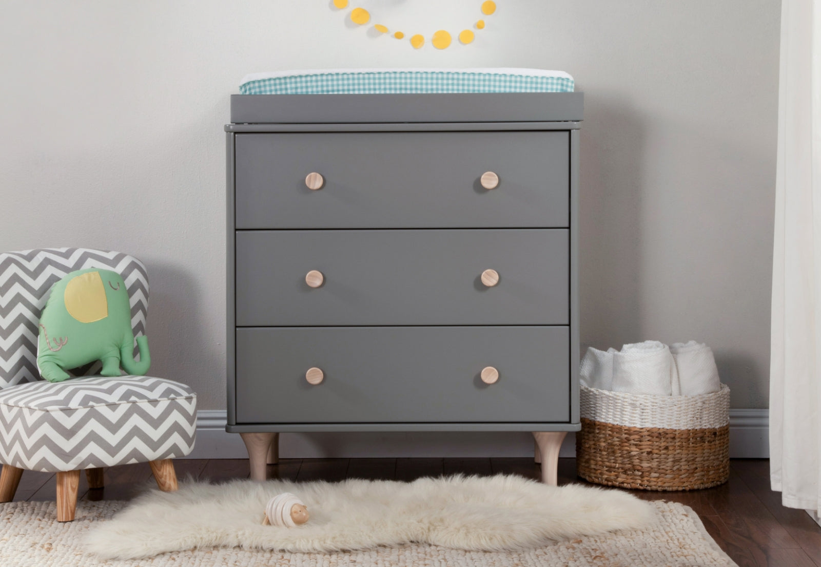 Modern nursery featuring a gray dresser with wooden knobs, accessorized with a plush elephant chair, a basket of linens, and a sheepskin rug on a textured carpet.